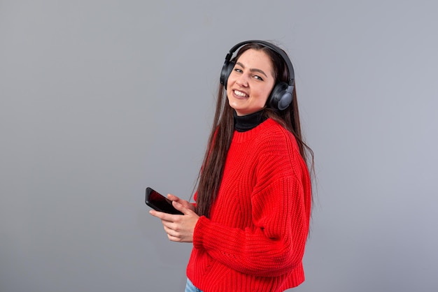 Positive girl brunette in red sweater listens to music on smartphone in headphones isolated on gray