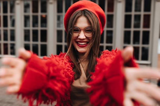 Positive girl in glasses laughs and stretches hands towards camera Lady dressed in red shaggy cardigan and beret posing against windows