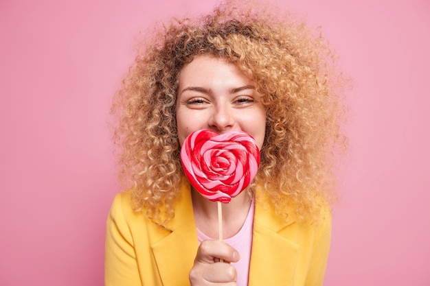 Positive good looking woman with curly bushy hair keeps heart shaped lollipop over mouth has sweet tooth likes sweets dressed in elegant outfit isolated over pink wall. Junk food concept