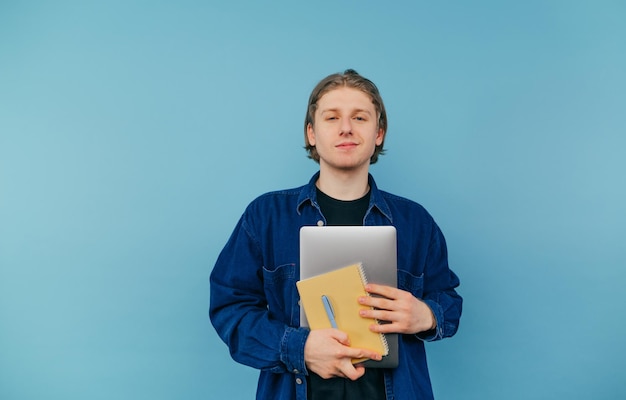 Positive guy in a blue shirt with a laptop and a notebook in his hand stands on a colored background