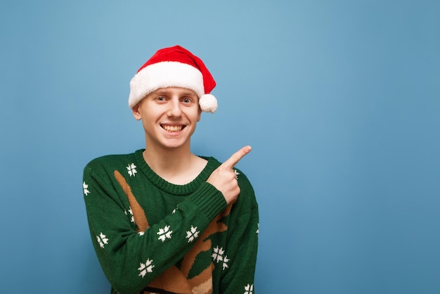 Positive guy in christmas sweater and hat santa stands against a blue background looks in camera