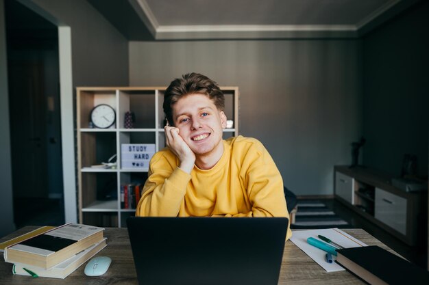 Positive guy student in casual clothes sitting at home in the bedroom at the table with books notebooks and laptop studying remotely looking at camera and smiling Quarantine training