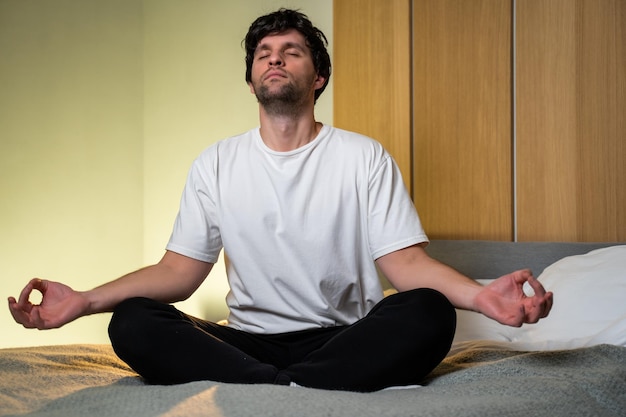 Positive guy in a white Tshirt and black pants sitting on the bed in the bedroom doing yoga in the evening sitting in the lotus position