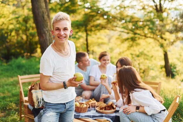 Positive guy with apple standing in front of people Group of young people have vacation outdoors in the forest Conception of weekend and friendship