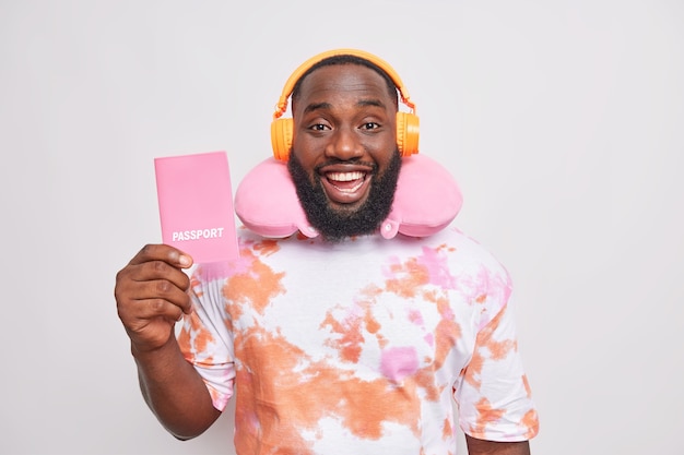 Positive handsome Afro American man holds passport listens music via headphones wears neck pillow for comfortable travel dressed in washed out t shirt isolated over white wall