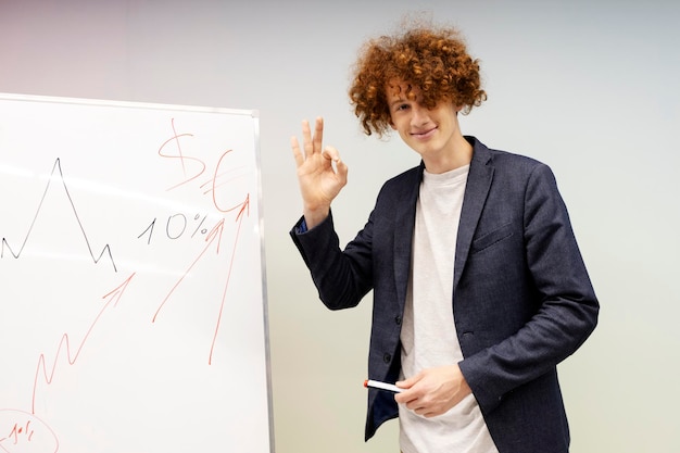 Positive handsome man showing ok gesture next to graphic drawn on flip chart Young businessman presenting successful business