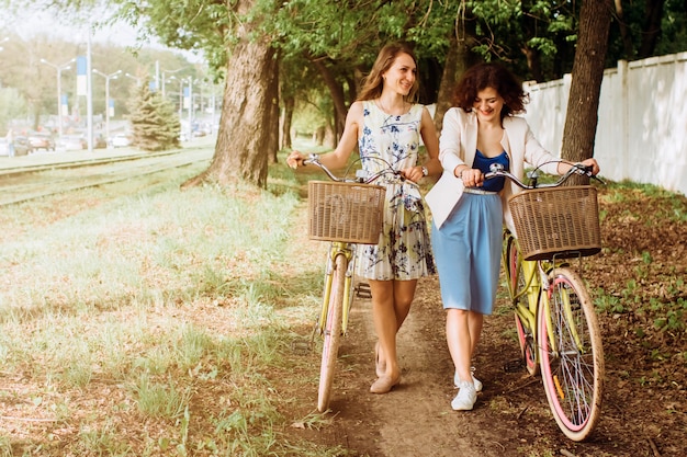 Positive and happy girls walking with bicycles on alley park, summer day. Female friends enjoying a walking down the street with their bikes.