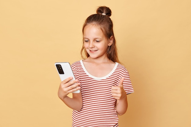 Positive happy smiling little brown haired girl wearing striped tshirt standing isolated over beige background using mobile phone showing like gesture enjoying internet content