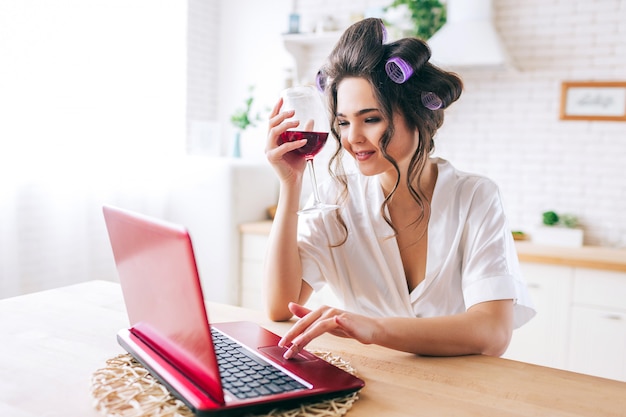 Positive happy young woman hold glass of red wine and work on laptop. Smiling. Home alone. Stand in kitchen. Morning daylight. Careless life of housekeeper.