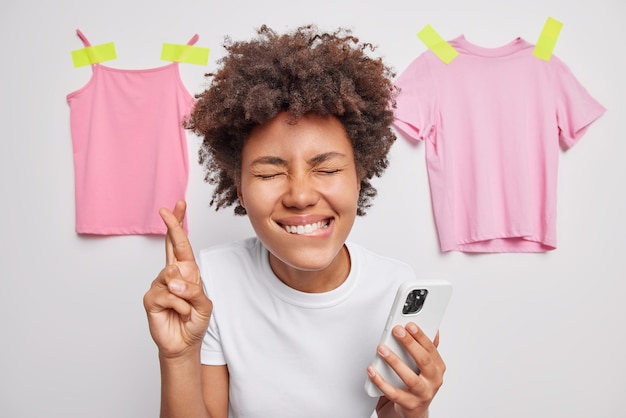 Positive hopeful woman bites lips and keeps fingers crossed believes in good luck closes eyes uses mobile phones dressed in casual t shirt poses against clothes background. Body language concept