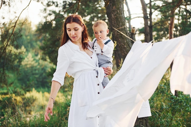 Positive housewife with hangs white clothes to dry Young mother with her little son is outdoors in the forest Beautiful sunshine