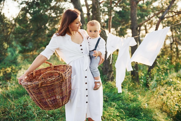 Positive housewife with hangs white clothes to dry Young mother with her little son is outdoors in the forest Beautiful sunshine