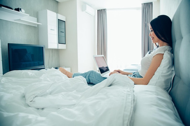 Positive lady sitting in the bed with a laptop on her laps and smiling while typing on a keyboard