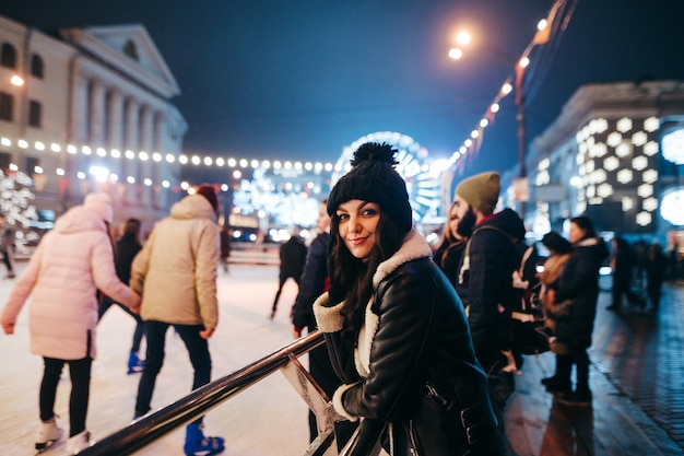 Positive lady in winter clothes stands at the rink at the Christmas street in evening and poses for the camera Portrait of a cute girl in a hat in the evening on Christmas fair