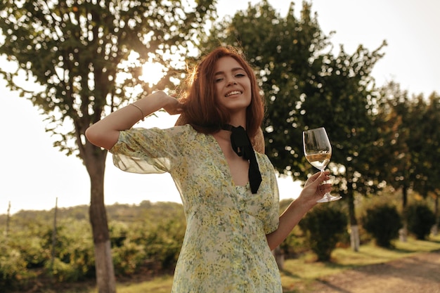 Positive lady with ginger hairstyle and bandage on neck in green clothes and hat smiling and holding with champagne outdoor