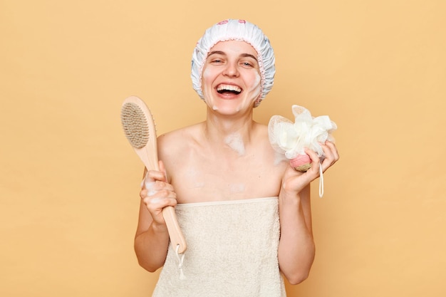 Photo positive laughing woman wearing shower cap standing isolated over beige background being in bath posing wrapped in towel holdingdry massage brush and washcloth