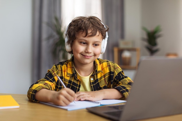 Positive little boy writing down exercise watching online lesson on laptop at home close up portrait