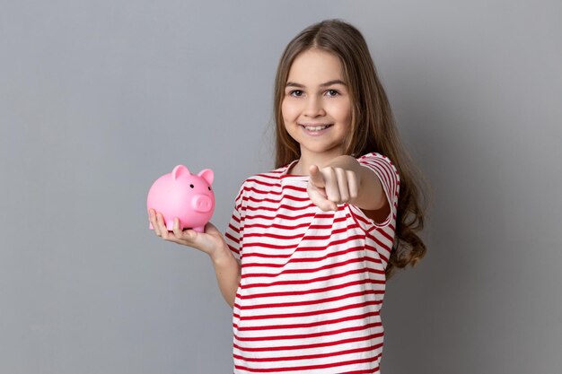 Positive little girl standing pointing at camera holding piggybank saving money banking