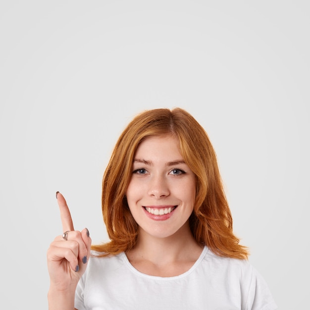 Positive lovely female with shining smile and red hair, points upwards with index finger, glad to present something, dressed in casual white t shirt, poses indoor alone.