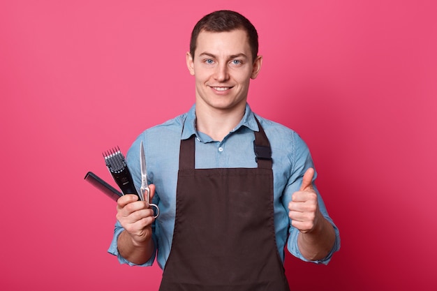 Positive male barber shows approval gesture, raises thumb, holds razor, scissors and comb, demostrates that he likes clients new hairstyle