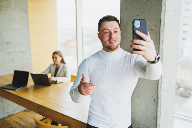 Positive male businessman in sweater smiling looking at phone standing in modern light office with phone working in modern company