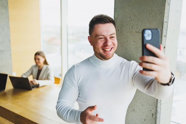 Positive male businessman in sweater smiling looking at phone standing in modern light office with phone working in modern company