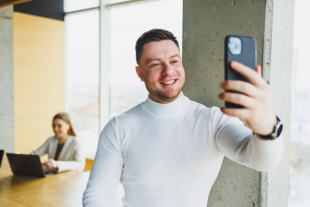 Positive male businessman in sweater smiling looking at phone standing in modern light office with phone working in modern company