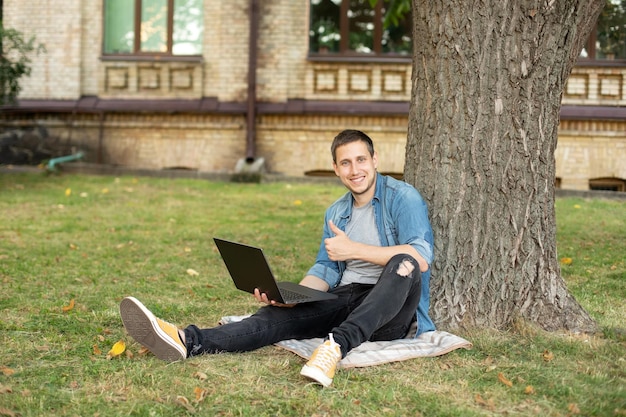 positive man hold laptop show thumb up on grass at university campus.