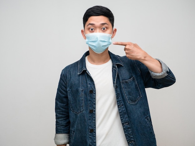 Positive man jeans shirt wearing medical mask point finger at his mask isolated