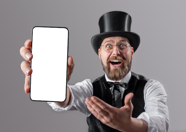 Positive mature aristocrat and suit and top hat showing modern smartphone with empty white screen while standing over grey studio background