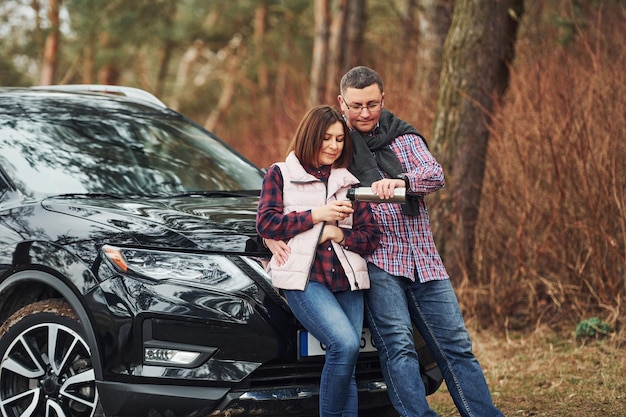 Positive mature couple standing near their car wit warm drink in hands Outdoors in forest