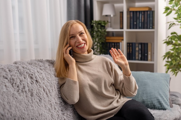 Positive mature woman in casual clothes sitting on couch and talking on modern smartphone Caucasian blonde enjoying mobile conversation during free time at cozy apartment