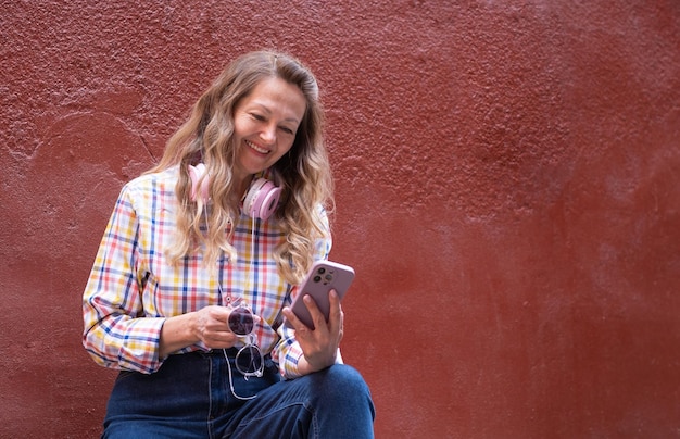 A positive mature woman with a smartphone in her hands sits against the background of a magenta color wall and listens to music