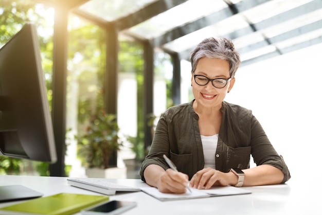 Positive middle aged woman working at home office taking notes