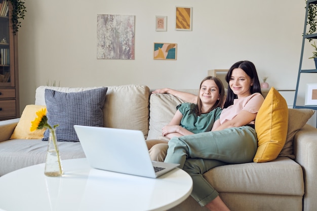 Positive mother and her teenage daughter sitting on sofa and watching movie on laptop during quarantine