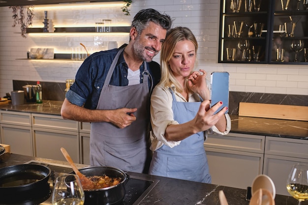 Positive multiracial couple in aprons standing near electric stove and taking self portrait on mobile phone while cooking food together at home