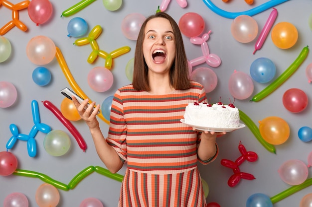 Positive overjoyed woman with brown hair wearing striped dress holding cake and smart phone in hands screaming with excitement standing against gray wall decorated with colorful balloons