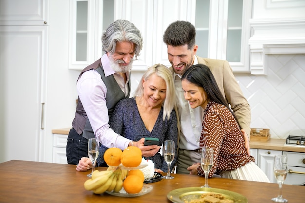 positive parents and smiling couple in formal wear looking at pictures or videos on smartphone