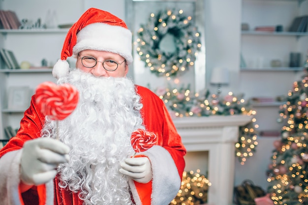 Positive and playful Santa Claus stands and looks. He holds two sweets. They have shape of heart.