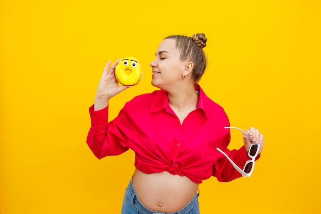 Positive pregnant woman eating donuts wearing pink shirt isolated on yellow background Happiness from pregnancy while expecting a child High quality photo Harmful food during pregnancy