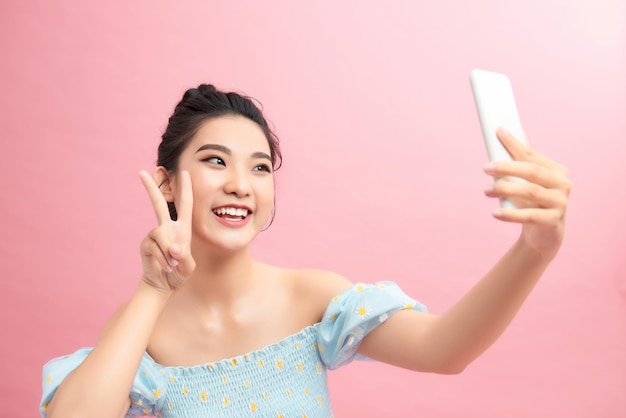 Positive pretty woman holding camera with extended arm showing peace sign taking selfie on smartphone with carefree smile