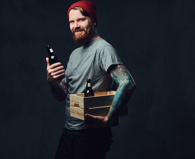 Positive redhead tattooed, bearded male holds a wooden box with beer bottles over grey background.