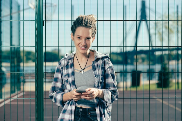 Positive relaxed young woman holding modern device and smiling while standing next to the chain link fence