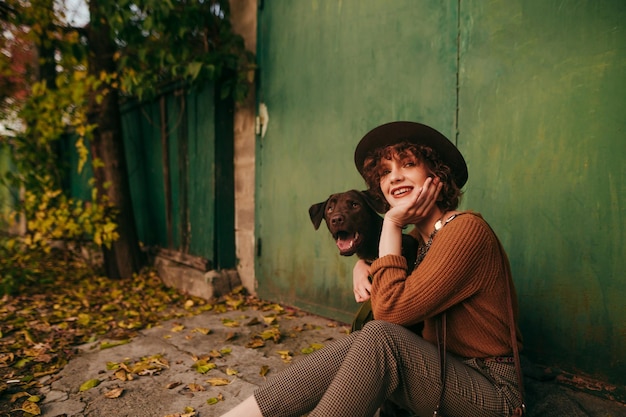 positive rural girl in vintage clothes and hat sitting with dog on the ground at country house