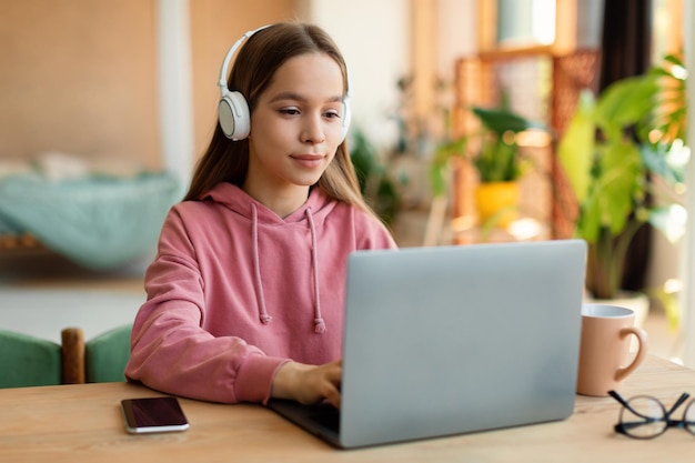 Positive schoolgirl in headphones watching video lesson remotely on laptop sitting at desk at home interior