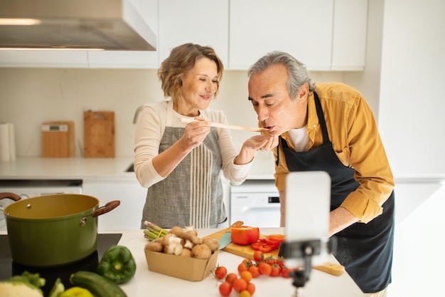 Positive senior spouses cooking healthy dinner and woman feeding husband streaming and using cellphon in kitchen