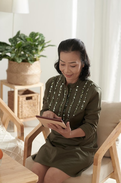 Positive senior woman checking social media application on tablet computer when spending time at home