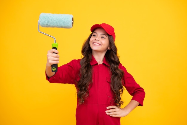 Positive and smiling builder in building uniform and cap Painter painting with paint roller Worker isolated on yellow background Kids renovation concept