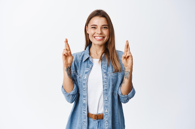 Positive smiling girl cross fingers for good luck and looking with hopeful face, making wish or praying, standing on white