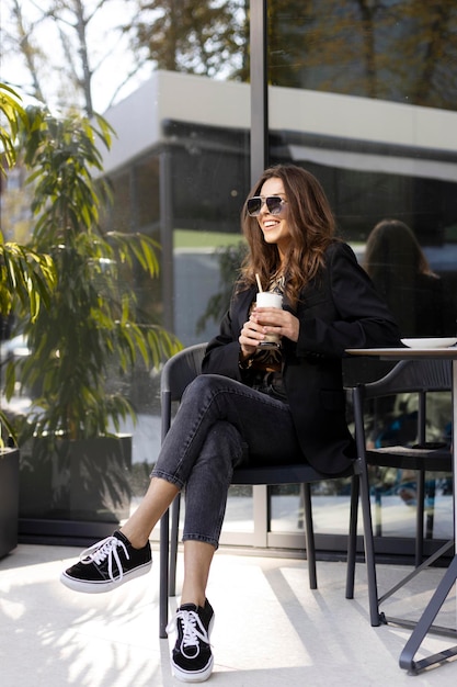 Positive smiling girl in sunglasses sits at a table on the street in a cafe Confident woman